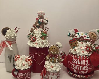 May include: A collection of holiday-themed decor featuring a white milk bottle, a red mug labeled Peppermint Kisses, a red canister with a heart, a small mug labeled Gingerbread Kisses, and a red teapot labeled Baking Spirits Bright, all topped with faux whipped cream and gingerbread figures.