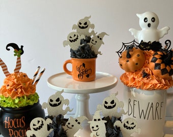 May include: Three orange mugs with black "boo" lettering and white ghost figures on top. The mugs are decorated with black and white frosting. The mugs are on a white surface with a white ghost figure and a black cat on a white mug in the background. The text "Beware" is on the white mug.