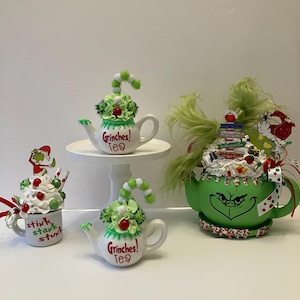 May include: A collection of whimsical Grinch-themed teacups and teapots. The teapots are white with green accents and the words "Grinches! Tea" in red. One teapot sits on a white pedestal. The teacups and teapots are decorated with green and white accents.