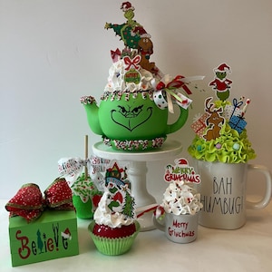 May include: A collection of Christmas-themed decorations featuring the Grinch. Includes a green teapot with a Grinch face, cupcakes, a mug that says "Bah Humbug!", and a wooden box that says "Believe!"
