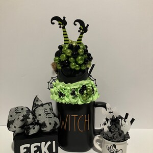 May include: A black mug labeled WITCH with green and black decorative spheres, witch legs, and spider accents. Beside it, a small white mug labeled BOO with ghost cutouts and a black block with a bow featuring the text EEK! in white lettering.