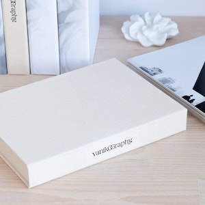 Puede incluir: Una caja rectangular blanca con el texto "vanikography" impreso en la parte delantera. La caja est&aacute; sobre una superficie de madera clara.