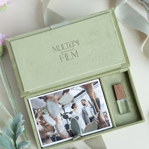 Op de afbeelding: Een saliegroene rechthoekige presentatiedoos met een scharnierend deksel met de tekst MULTANI PRODUCTIONS FILM. Binnenin is een rechthoekige foto van een koppel in een kas geplaatst naast een doorzichtige USB-stick met een houten dop.