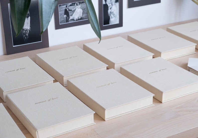 Puede incluir: Una fila de ocho cajas beige con el texto "moments of love" impreso en dorado en la parte delantera. Las cajas est&aacute;n dispuestas sobre una superficie de madera clara.