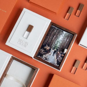 Puede incluir: Una caja de presentación blanca y de madera oscura que contiene una fotografía de una boda en un bosque, con el texto "MEGAN GIELOW" grabado en la tapa. Varios frascos pequeños de vidrio transparente con tapas de madera están dispuestos.