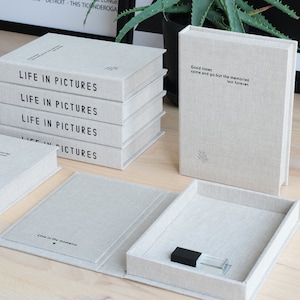Puede incluir: Un conjunto de cinco álbumes de fotos cubiertos de lino con el título "Life in Pictures" impreso en la parte delantera. Los álbumes están apilados uno encima del otro. Una caja más pequeña cubierta de lino con una cita sobre los recuerdos está abierta y tiene una pequeña botella transparente en su interior.