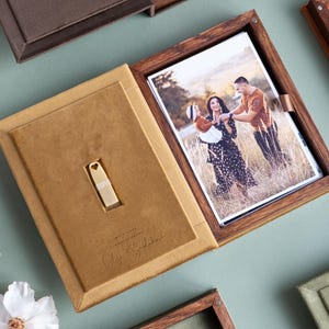 Può includere: Una scatola fotografica in legno con interno in pelle scamosciata marrone chiaro, contenente una fotografia e una chiavetta USB. La scatola è aperta, rivelando un ritratto di famiglia. Il testo "With Love Photography" e una firma sono visibili.