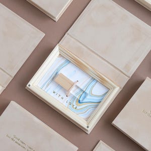 May include: Wooden box with a light-colored finish, open to reveal a framed print with blue and gold abstract art. A wooden USB drive is inside. The text "WITH LOVE" is visible. Other closed boxes are scattered around.