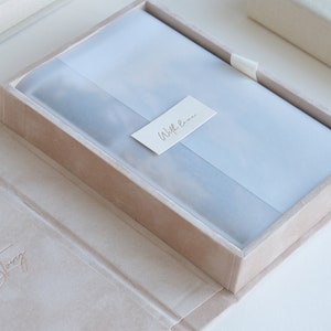 Puede incluir: Una caja de terciopelo marr&oacute;n claro con un forro de sat&eacute;n blanco. La caja est&aacute; abierta y el forro es visible. La caja tiene las palabras "Our Love Story" grabadas en la parte delantera. La caja est&aacute; cerrada con una cinta blanca. La caja est&aacute; sentada sobre una superficie blanca.