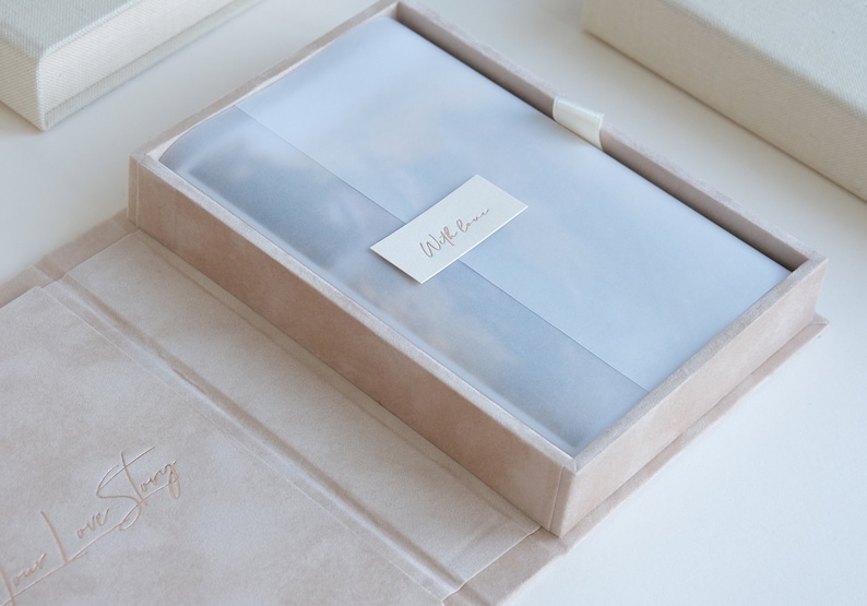 Puede incluir: Una caja de terciopelo marr&oacute;n claro con un forro de sat&eacute;n blanco. La caja est&aacute; abierta y el forro es visible. La caja tiene las palabras "Our Love Story" grabadas en la parte delantera. La caja est&aacute; cerrada con una cinta blanca. La caja est&aacute; sentada sobre una superficie blanca.