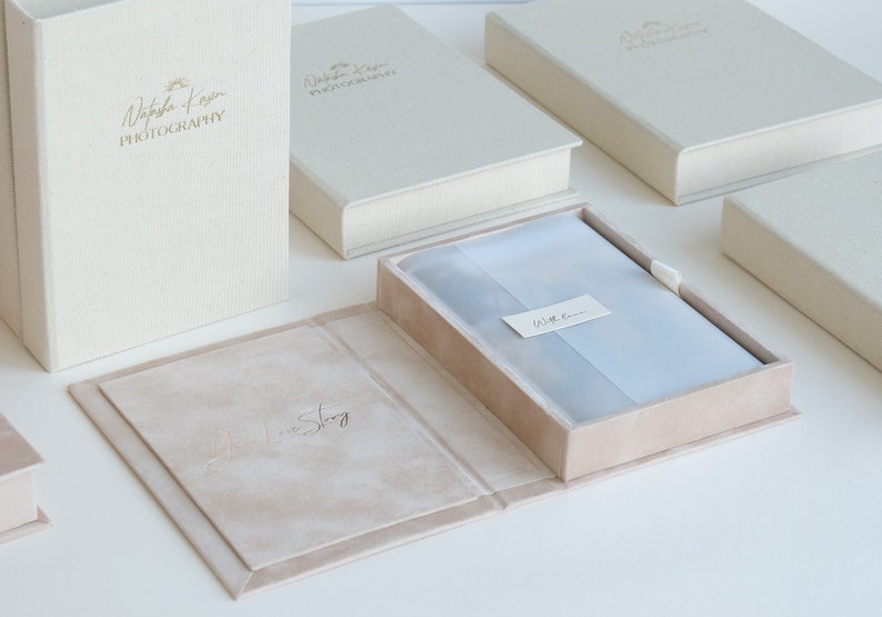 Puede incluir: Un conjunto de cajas beige y crema con un logotipo en relieve dorado que dice "Natasha Kayin Photography". Las cajas est&aacute;n hechas de una tela texturizada y tienen un interior de terciopelo. Las cajas est&aacute;n dise&ntilde;adas para guardar fotograf&iacute;as y otros recuerdos.