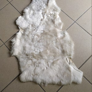 May include: A white sheepskin rug with a soft, fluffy texture. The rug is laid out flat on a tiled floor.