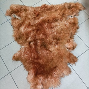 May include: A brown sheepskin rug with a soft, fluffy texture. The rug is laid out flat on a tiled floor.