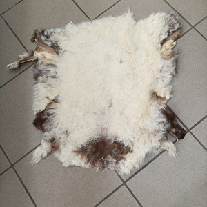 May include: A white sheepskin rug with brown patches. The rug is lying on a tiled floor.