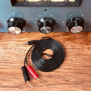 May include: Close-up of a vintage audio equipment panel with a VU meter and control knobs. A black cable with red and black connectors and a 1/4 inch jack is coiled on a wooden surface. The meter needle points to the right.