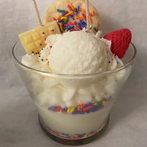 May include: A white candle shaped like a sundae with a white chocolate bar, a white cookie with colorful sprinkles, and a red strawberry on top. The candle is in a clear glass jar with a colorful base.