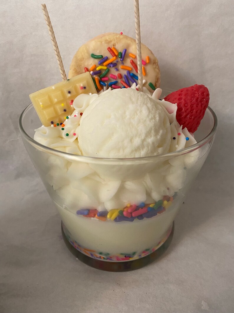 May include: A white candle shaped like a sundae with a white chocolate bar, a white cookie with colorful sprinkles, and a red strawberry on top. The candle is in a clear glass jar with a colorful base.