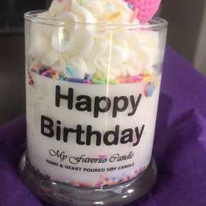 May include: A white soy candle in a clear glass jar with a white frosting design, sprinkles, and a pink strawberry on top. The label reads "Happy Birthday" and "My Favorite Candle, Hand & Heart Poured Soy Candle".