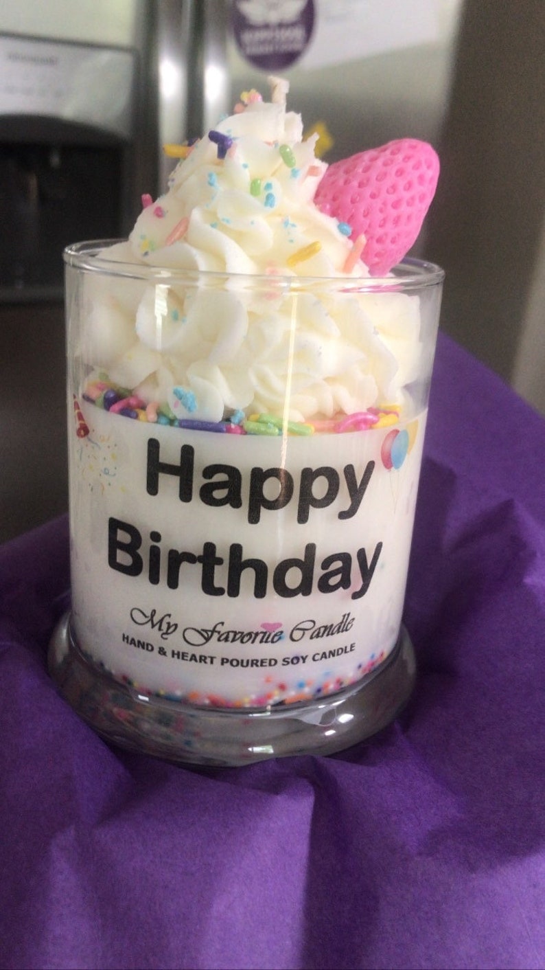 May include: A white soy candle in a clear glass jar with a white frosting design, sprinkles, and a pink strawberry on top. The label reads "Happy Birthday" and "My Favorite Candle, Hand & Heart Poured Soy Candle".