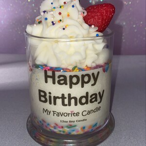 May include: A white soy candle in a clear glass jar with a white frosting design, colorful sprinkles, and a red strawberry on top. The jar has a gold label that reads "Happy Birthday My Favorite Candle 12oz Soy Candle".