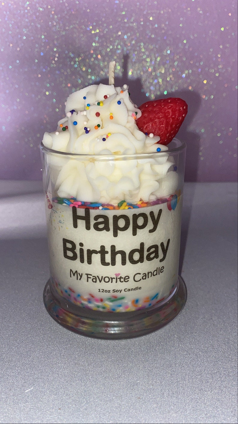May include: A white soy candle in a clear glass jar with a white frosting design, colorful sprinkles, and a red strawberry on top. The jar has a gold label that reads "Happy Birthday My Favorite Candle 12oz Soy Candle".