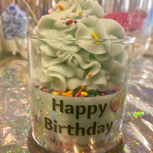 May include: A green cupcake candle in a clear glass jar with sprinkles and the words "Happy Birthday" on the label.