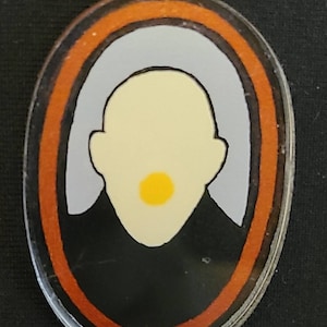 May include: A black and white oval pin with a stylized image of a person's head. The person has a yellow circle in the center of their face.
