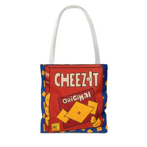 May include: A red and blue tote bag with a white handle. The bag features a cartoon image of a box of Cheez-It crackers. The box says "Cheez-It Original" and "100% Real Cheese".