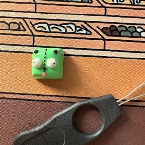 May include: A green and white keycap with a cartoon face and black eyes. The keycap is sitting on a brown and orange surface with a black tool.