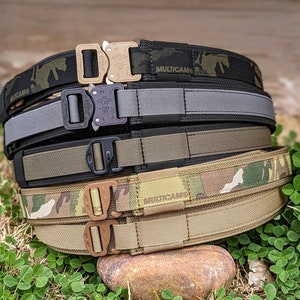 Könnte beinhalten: Ein Stapel von vier taktischen Gürteln in verschiedenen Tarnmustern, darunter Multicam und ein einfarbiger schwarzer Gürtel. Die Gürtel haben Metallschnallen und sind aus strapazierfähigem Nylon gefertigt.