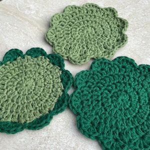 Op de afbeelding: Drie handgemaakte gehaakte onderzetters in groentinten. Elke onderzetter heeft een geschulpte rand, met verschillende tinten groen garen gebruikt om een ​​gestructureerd, decoratief ontwerp te creëren. De onderzetters zijn cirkelvormig.