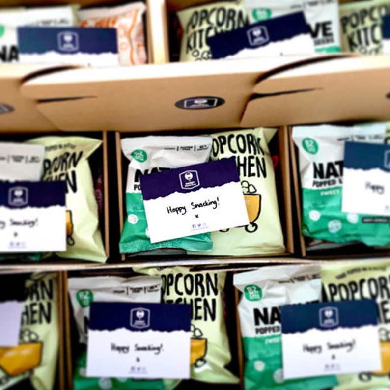 May include: A close-up of several boxes filled with popcorn bags. The boxes are labeled with the text "Popcorn Kitchen" and "Happy Snacking!" The bags are various colors, including green, blue, and white.