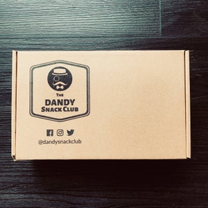 May include: A brown cardboard box with a logo that says "The Dandy Snack Club" and a cartoon image of a man with a monocle and a top hat. The logo is in a black circle with a black border. The box also has social media icons and the text "@dandysnackclub".