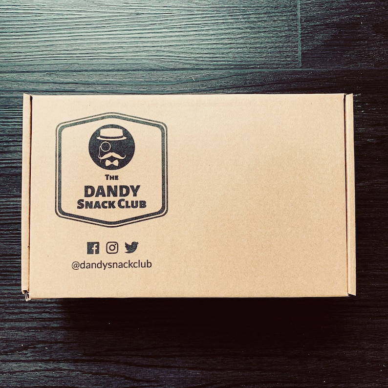 May include: A brown cardboard box with a logo that says "The Dandy Snack Club" and a cartoon image of a man with a monocle and a top hat. The logo is in a black circle with a black border. The box also has social media icons and the text "@dandysnackclub".