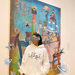 以下が含まれることがあります： 大きなカラフルな絵画の前に、グレーの文字が入った白いパーカーを着た人。絵画は、さまざまな人物やオブジェクトが描かれた青い背景を持っています。装飾的な星と惑星が画像に追加されています。