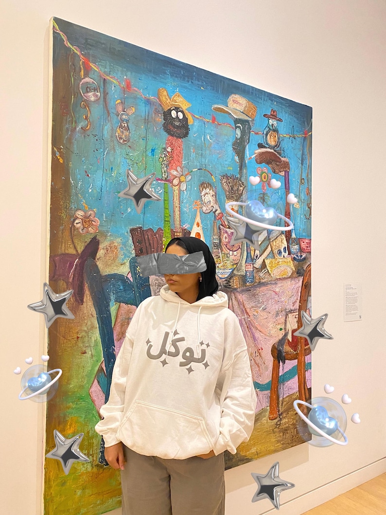以下が含まれることがあります： 大きなカラフルな絵画の前に、グレーの文字が入った白いパーカーを着た人。絵画は、さまざまな人物やオブジェクトが描かれた青い背景を持っています。装飾的な星と惑星が画像に追加されています。