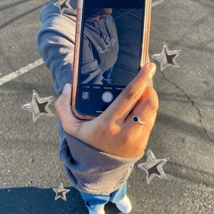 以下が含まれることがあります： 赤いケースに入ったスマートフォンには、アラビア語のテキストが入ったグレーのパーカーを着た人の写真が表示されています。その人は青いジーンズと白いスニーカーを履いています。銀色の星の装飾が携帯電話を囲んでいます。
