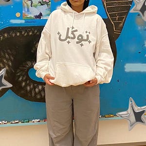 以下が含まれることがあります： グレーの文字が入った白いパーカーとグレーのパンツを着た人。パーカーにはドローストリングフードとカンガルーポケットが付いています。パンツは明るいグレーです。白と黒のスニーカーを履いています。