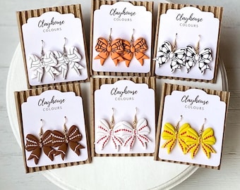 May include: Six pairs of handmade polymer clay earrings shaped like bows with different colour and pattern combinations. The earrings are displayed on cards with the text "Clayhouse Colours".