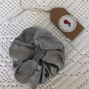 May include: A grey fabric scrunchie with a subtle striped pattern. The scrunchie is tied in a knot and is lying on a white crocheted surface.