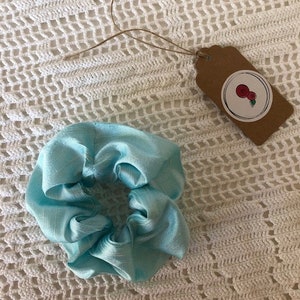 May include: A light blue satin scrunchie hair tie. The scrunchie is made of soft, silky fabric and is perfect for adding a touch of elegance to any hairstyle.