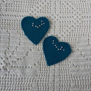 May include: Two teal felt heart-shaped ornaments with small white beads arranged in a curved line. The hearts are placed on a white crocheted surface, creating a contrast in texture and color. The ornaments are likely handmade.