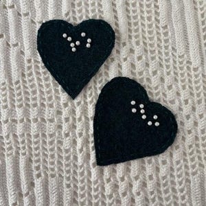 May include: Two dark green heart-shaped fabric patches featuring a constellation pattern made of small white beads. Each heart has a visible stitched border in a matching dark green thread, creating a textured and detailed appearance on the fabric.