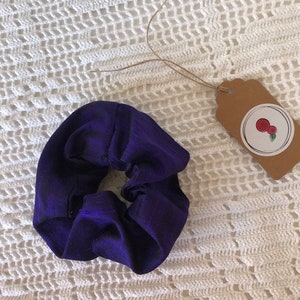 May include: A dark purple scrunchie made of fabric. The scrunchie is lying on a white crocheted surface.