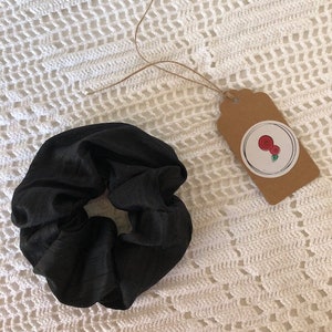 May include: Black scrunchie made of silky fabric. The scrunchie is tied in a knot and is lying on a white crocheted surface.