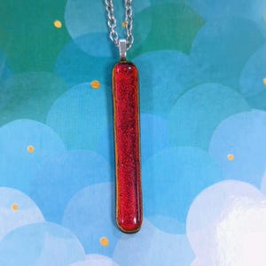 May include: A red rectangular pendant necklace with a silver chain. The pendant has a textured, glittery appearance. The necklace is displayed against a blue and green background with white cloud shapes and yellow dots.