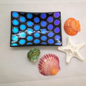 May include: A black rectangular dish with blue polka dots, alongside an orange seashell, a white starfish, a green seashell, and a pink seashell. The dish has a glossy finish, and the seashells are in various shapes.