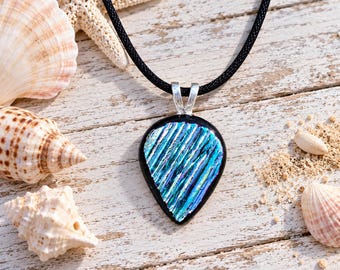 May include: A teardrop-shaped pendant necklace featuring a black glass base with a textured, iridescent blue and purple striped pattern. The pendant is attached to a simple silver-toned metal bail and hangs from a smooth black cord necklace.
