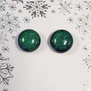 May include: A pair of round stud earrings featuring a dark green glitter finish with a smooth, domed surface. The earrings are displayed on a white card decorated with silver snowflake patterns along the edges, providing a festive presentation.