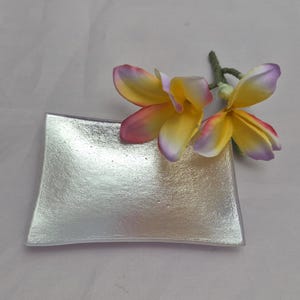 May include: A silver-colored, rectangular dish with slightly curved edges. Two yellow and pink flowers with purple tips rest on the dish. The dish has a textured, metallic appearance, and is set against a light background.